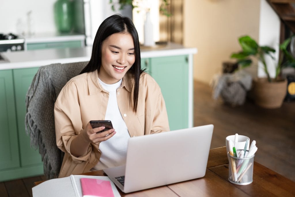 Smiling Asian Woman Using Laptop At Home. Girl Holding Smartphone And Watching Webinar On Computer